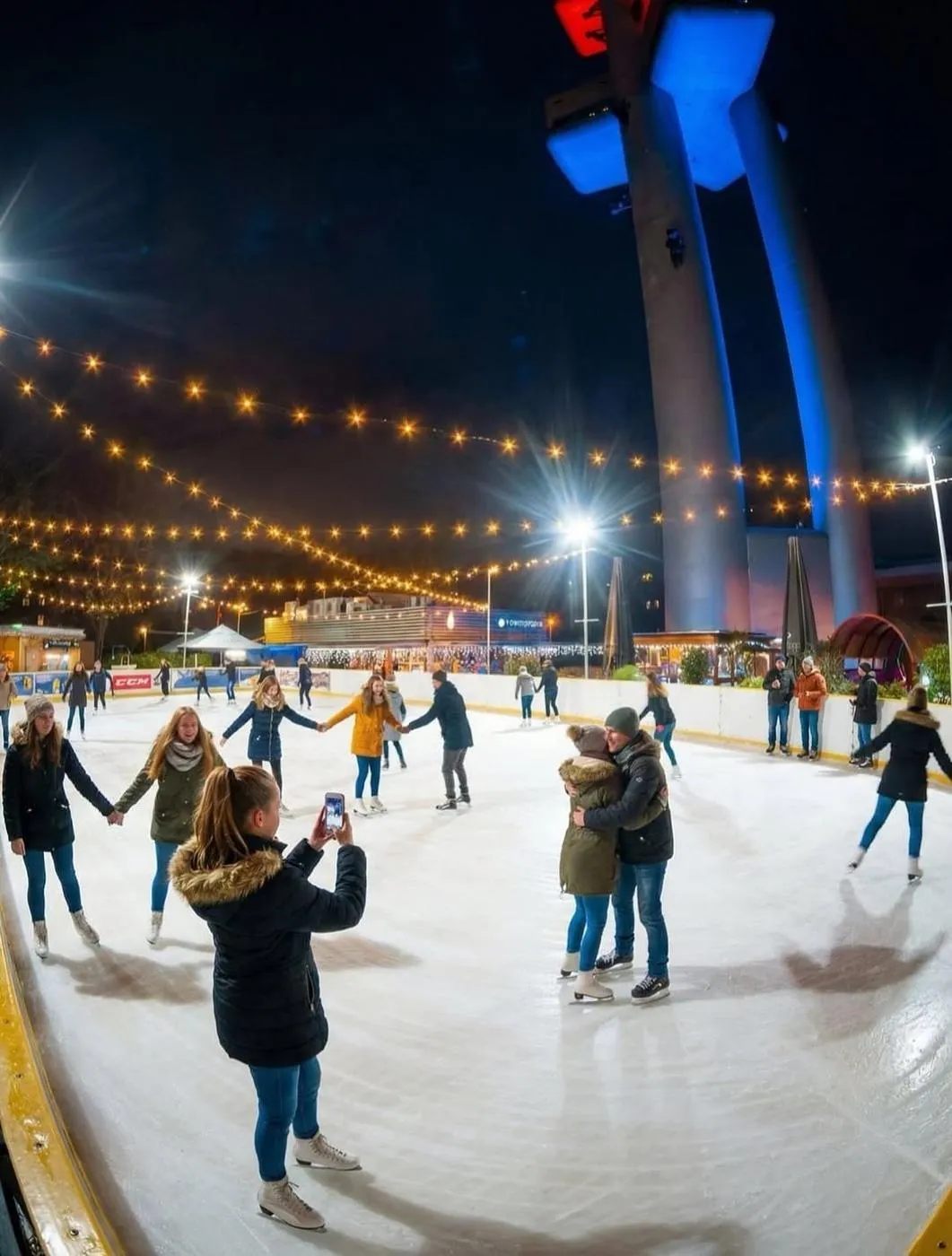 Skaters on the Pod Žižkovskou věží rink at night, with the blue-lit television tower above and strings of festoon lights over the ice. — Kluziště Pod Žižkovskou věží (Praha 3)