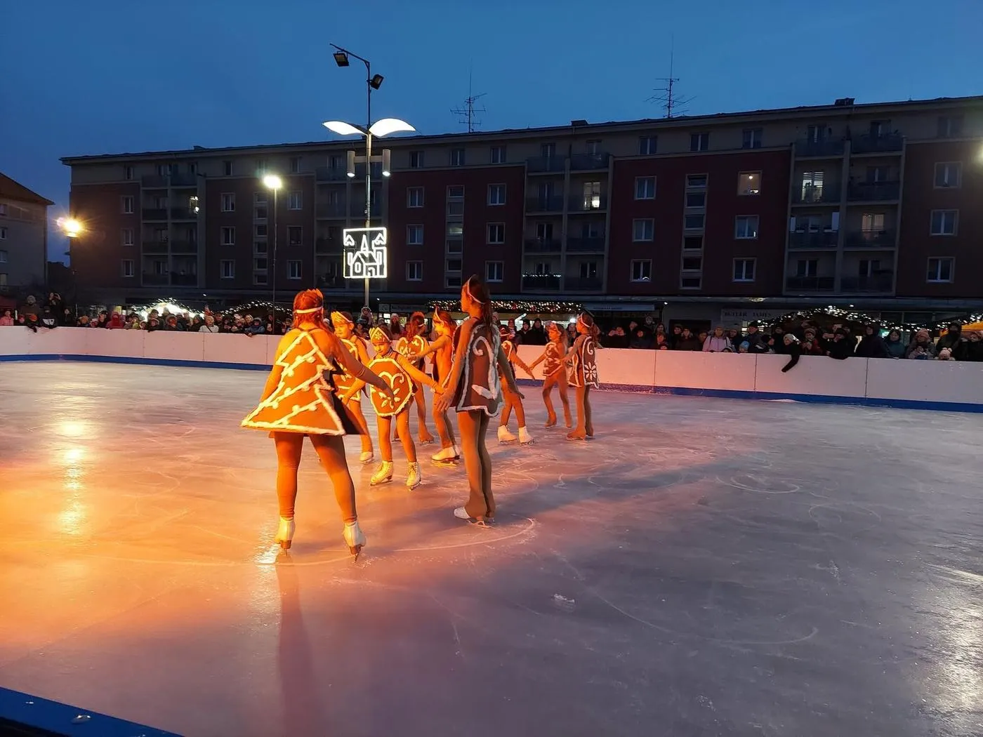 Performers in orange costumes skating a show on the lit ice of the Příbram rink at dusk, with an apartment block behind the boards. — Kluziště Příbram