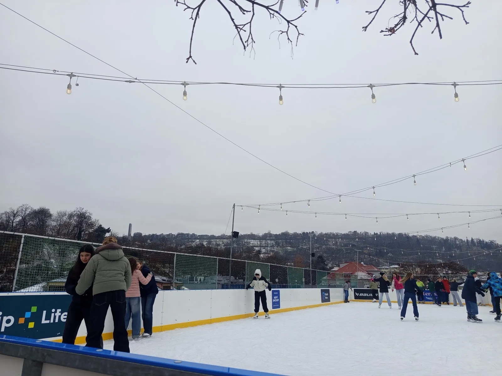 The rooftop ice with strings of lights over the boards, a few skaters at the far end and the hills of Prague beyond. — Terasa Smíchov Ice Rink