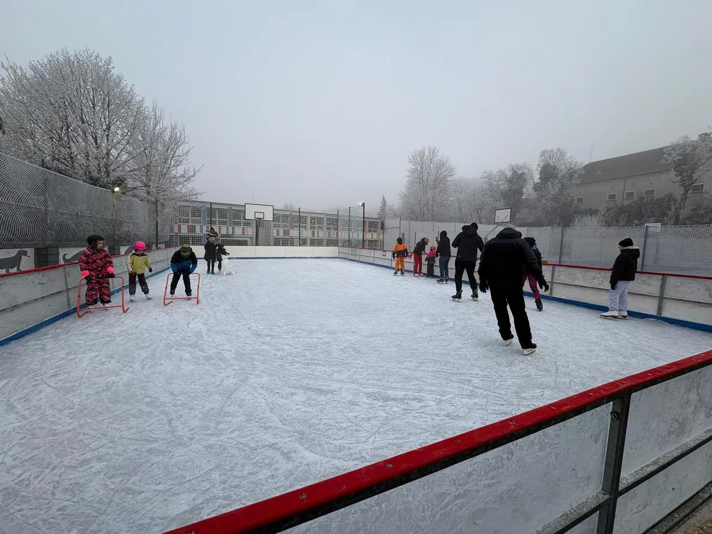 Children and adults skating on the fenced rink at Uhříněves on a misty winter day, with hockey goals on the ice, a red rail and school buildings behind. — Kluziště Uhříněves (Praha Uhříněves)