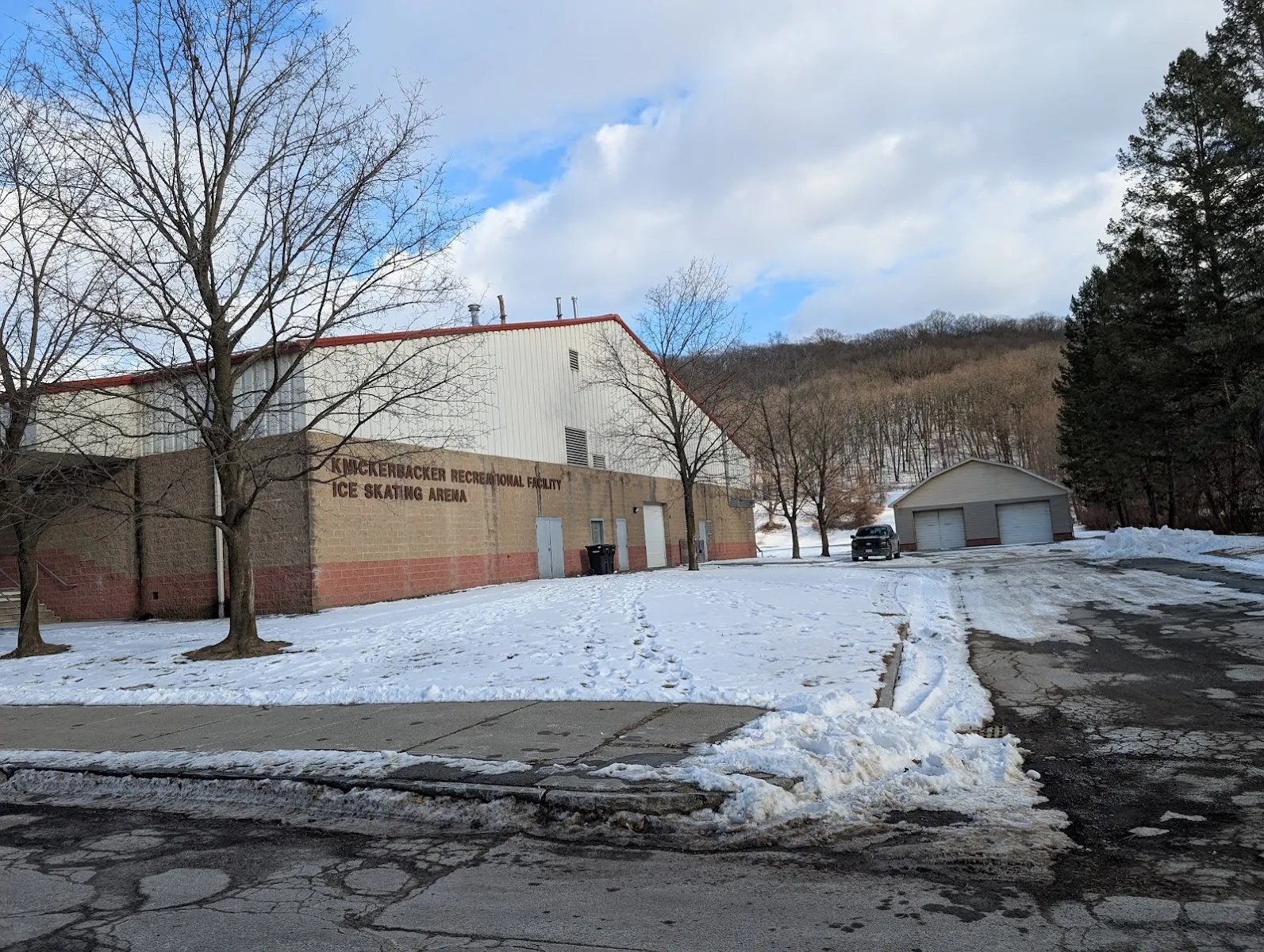 Knickerbacker Recreational Facility Ice Skating Arena building in winter, tan brick base with red-trimmed white metal siding and "KNICKERBACKER RECREATIONAL FACILITY ICE SKATING ARENA" lettering, snow-covered lawn and parking area, wooded hillside behind. — Knickerbacker Ice Arena