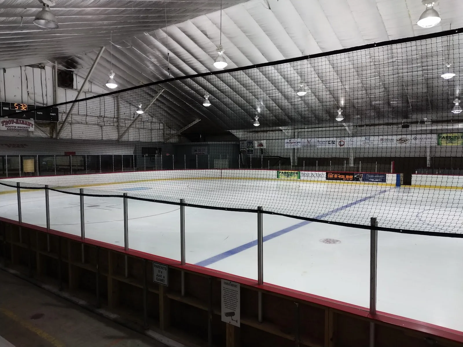 An empty ice sheet inside Knickerbacker Ice Arena, with dasher boards and glass along the near side and a steel-framed roof overhead. — Knickerbacker Ice Arena