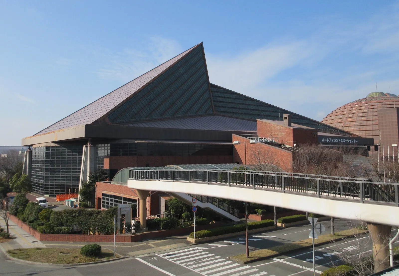 歩道橋から見た、ピラミッド形の屋根の神戸ポートアイランドスポーツセンターの建物。右側の壁にセンターの名前があります — Kobe Port Island Sports Center Ice Rink