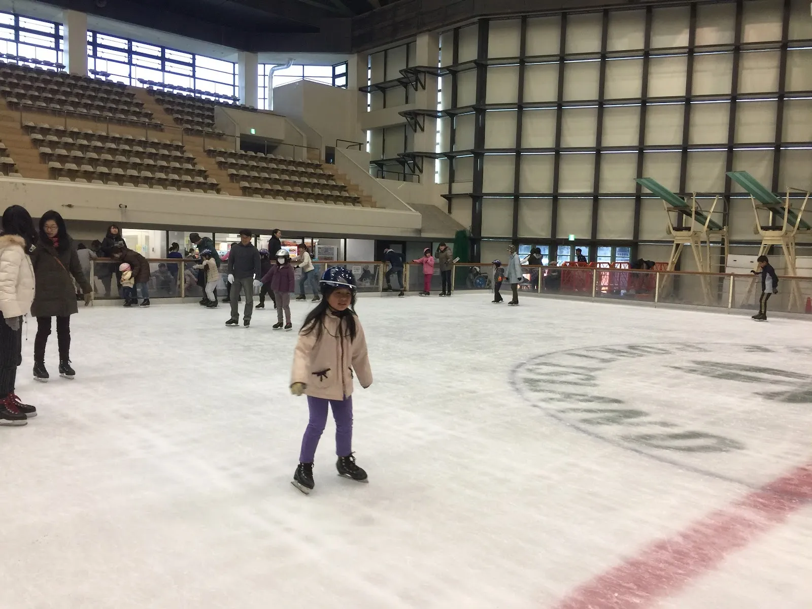神戸ポートアイランドスポーツセンターの一般滑走の様子。氷面全体にスケーターが広がり、フェンスの奥に客席があります。 — Kobe Port Island Sports Center Ice Rink