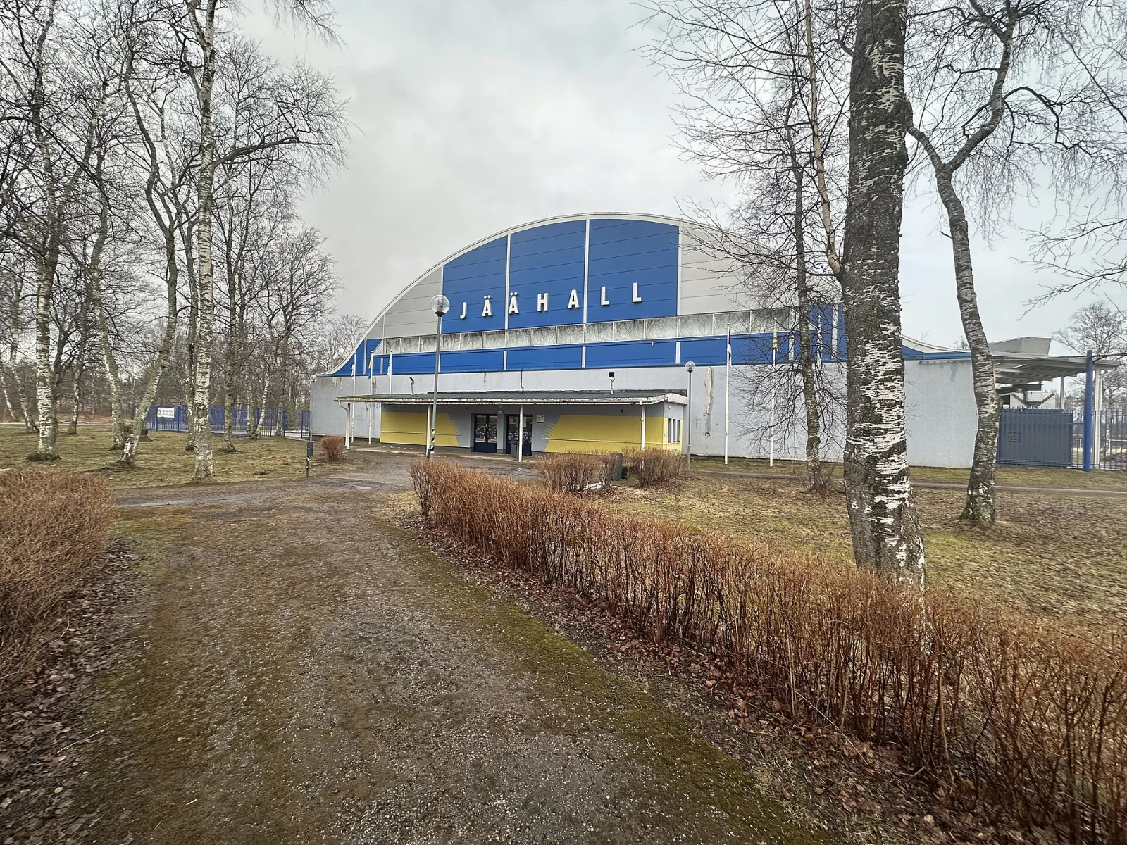 The Kohtla-Järve ice hall seen through the birches, with its arched blue roof front and JÄÄHALL sign — Kohtla-Järve jäähall
