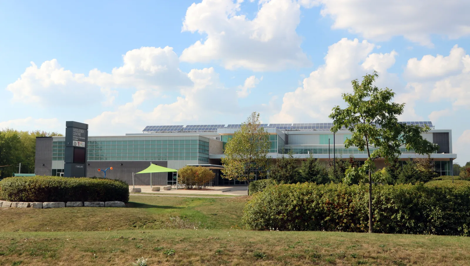 The Komoka Wellness Centre's long, low front seen across the lawn and young trees, under a partly cloudy sky. — Komoka Wellness Centre