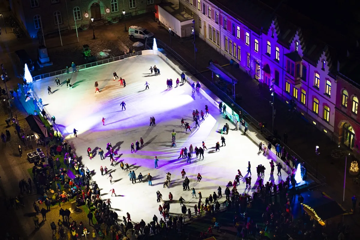 Aerial view of the Kompis ice rink on the town square in Kristiansand at night, with skaters on the lit ice and the historic buildings around the square. — Isbanen Kompis