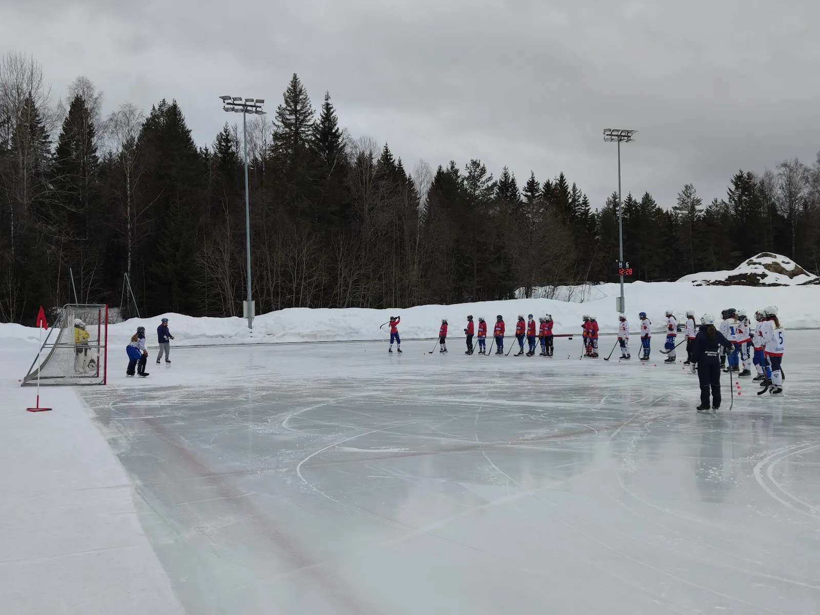 The outdoor ice pad in winter with its rink lines and the trees behind, the far end with hockey players at distance and a goal. — Konnerud kunstisbane