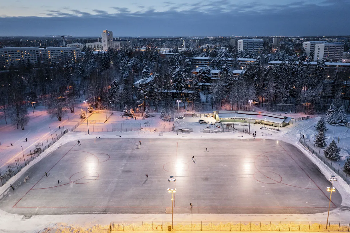Aerial view of Kontula sports park with the artificial ice rink in the foreground and the snowy local sports field behind it. — Kontulan liikuntapuisto – luistelukaukalo