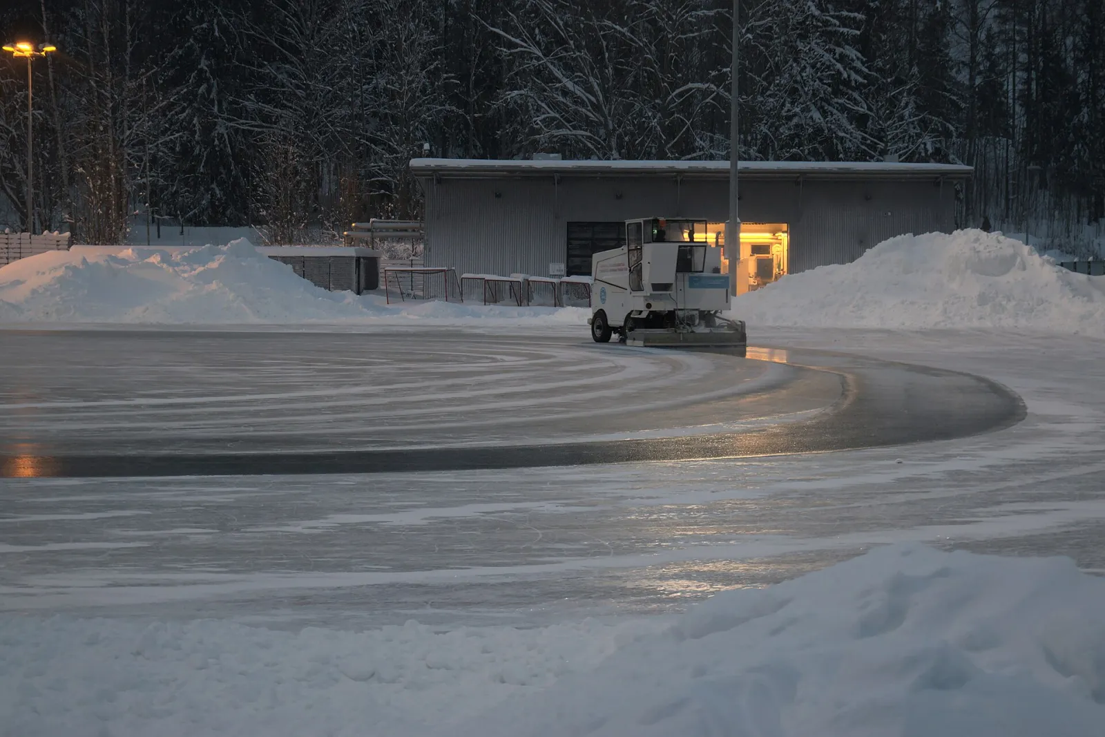 A machine resurfacing the artificial ice at Kontula sports park in winter, with the snow-banked rink edge and the service building behind. — Kontulan liikuntapuisto – luistelukaukalo