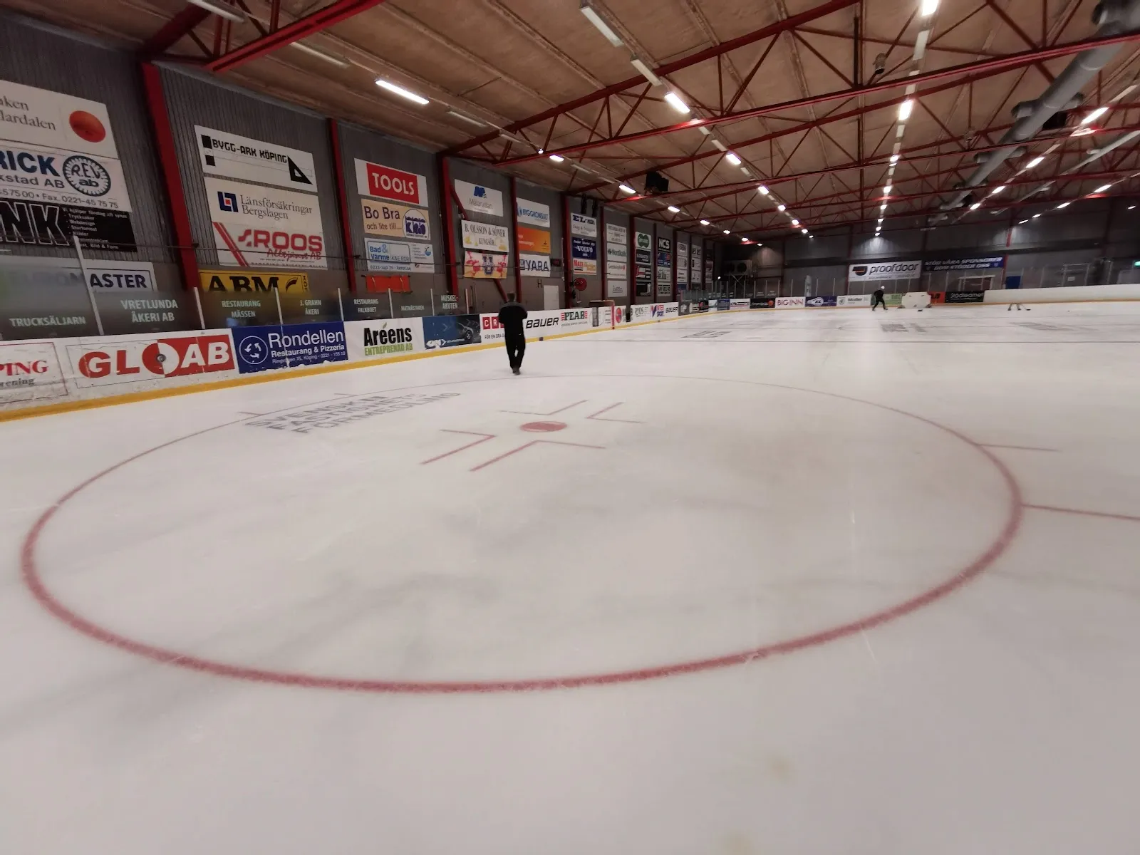 Den tomma isen i Köpings ishall med sin mittcirkel och sponsorsargar under de röda takbalkarna – Köpings ishall