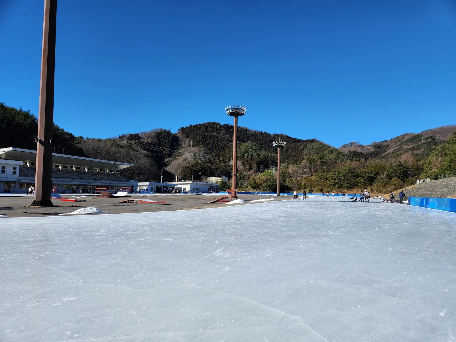 The oval's clean ice under a clear blue sky, with floodlight masts, the low grandstand and clubhouse buildings and wooded hills on the far side of the track. — Koriyama Skating Rink (Bandai Atami Sports Park)
