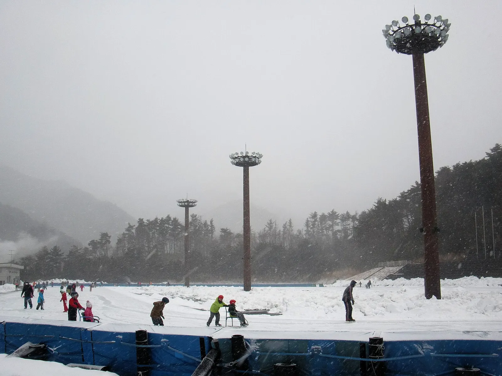The oval's snow-edged ice seen across the track from the boards, with children skating and sledding in the distance and a tall floodlight mast beside the course. — Koriyama Skating Rink (Bandai Atami Sports Park)