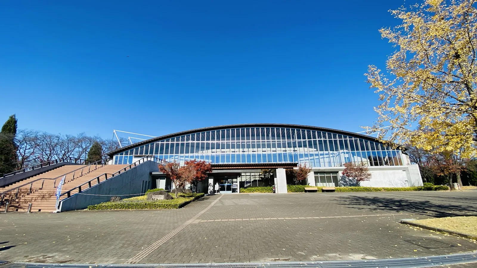 晴れた空の下の、甲府にある小瀬スポーツ公園アイスアリーナの、曲線を描くガラス張りの建物。舗装された前庭があり、左にスロープがあります — Kose Sports Park Ice Arena