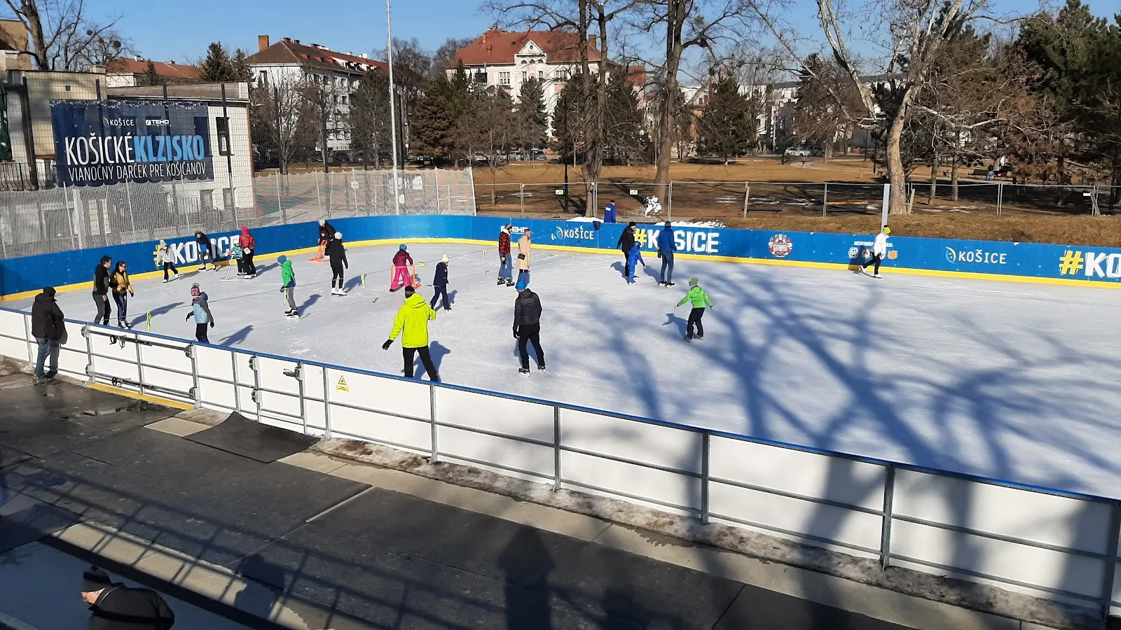 Skaters spread across the outdoor Košice rink in winter sun, with blue boards, trees and old buildings of Mestský park behind. — Košické klzisko