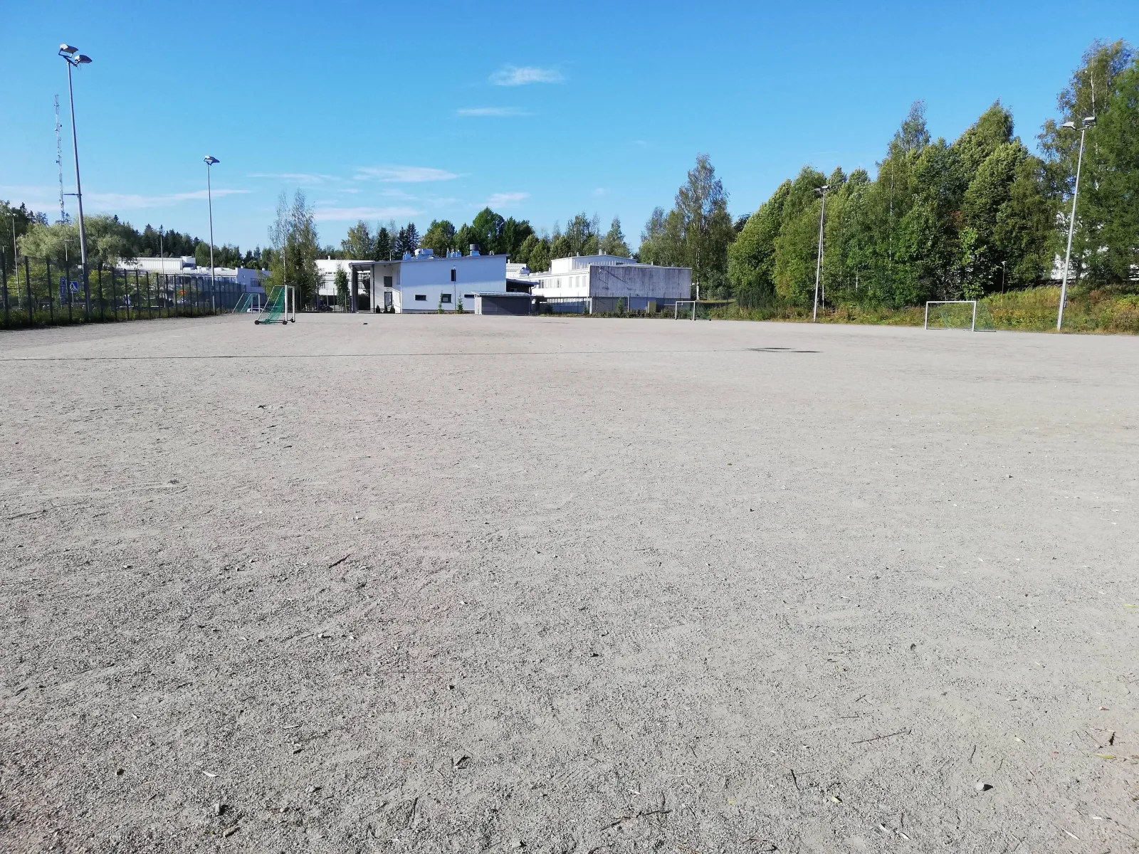The wide gravel field at Koulupuisto in Kuopio, which serves as a skating field in winter, seen in summer with floodlight poles and school buildings in the distance. — Koulupuiston luistelukenttä