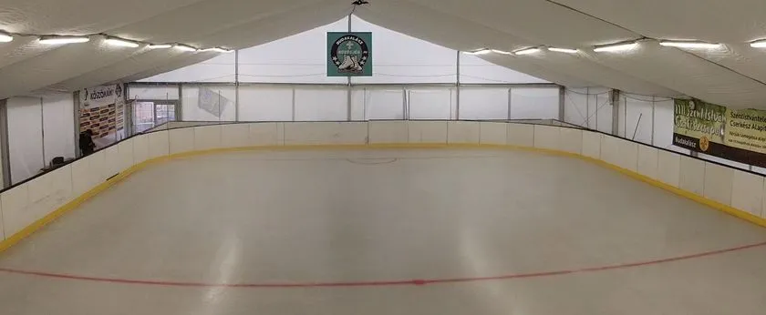 The empty boarded ice sheet inside the white tent hall of the Közösjég rink, with a red centre line, fluorescent strip lights under the tent roof and a club banner on the far wall. — Közösjég Budakalász