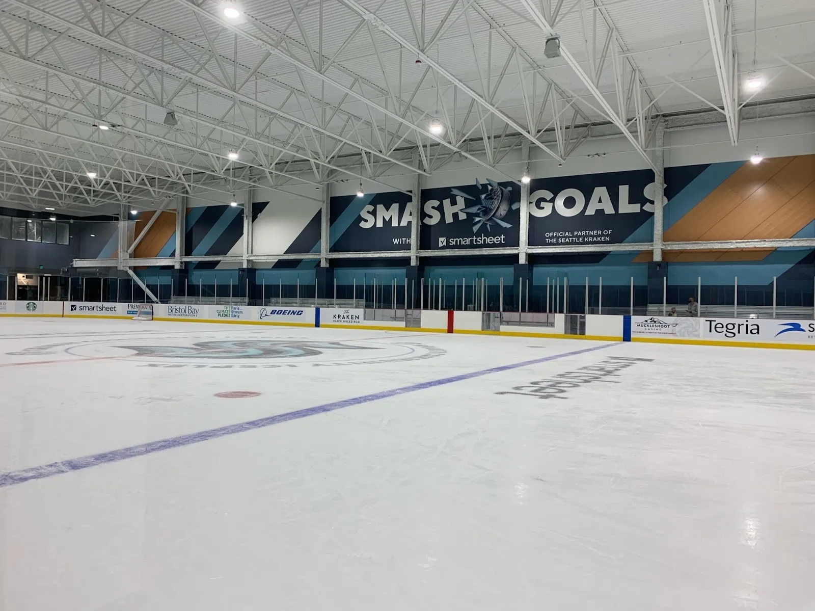 La pista vacía junto a las vallas del extremo, con una pared de banderines de Smash Goals sobre el vidrio y un techo con estructura de acero arriba. — Kraken Community Iceplex