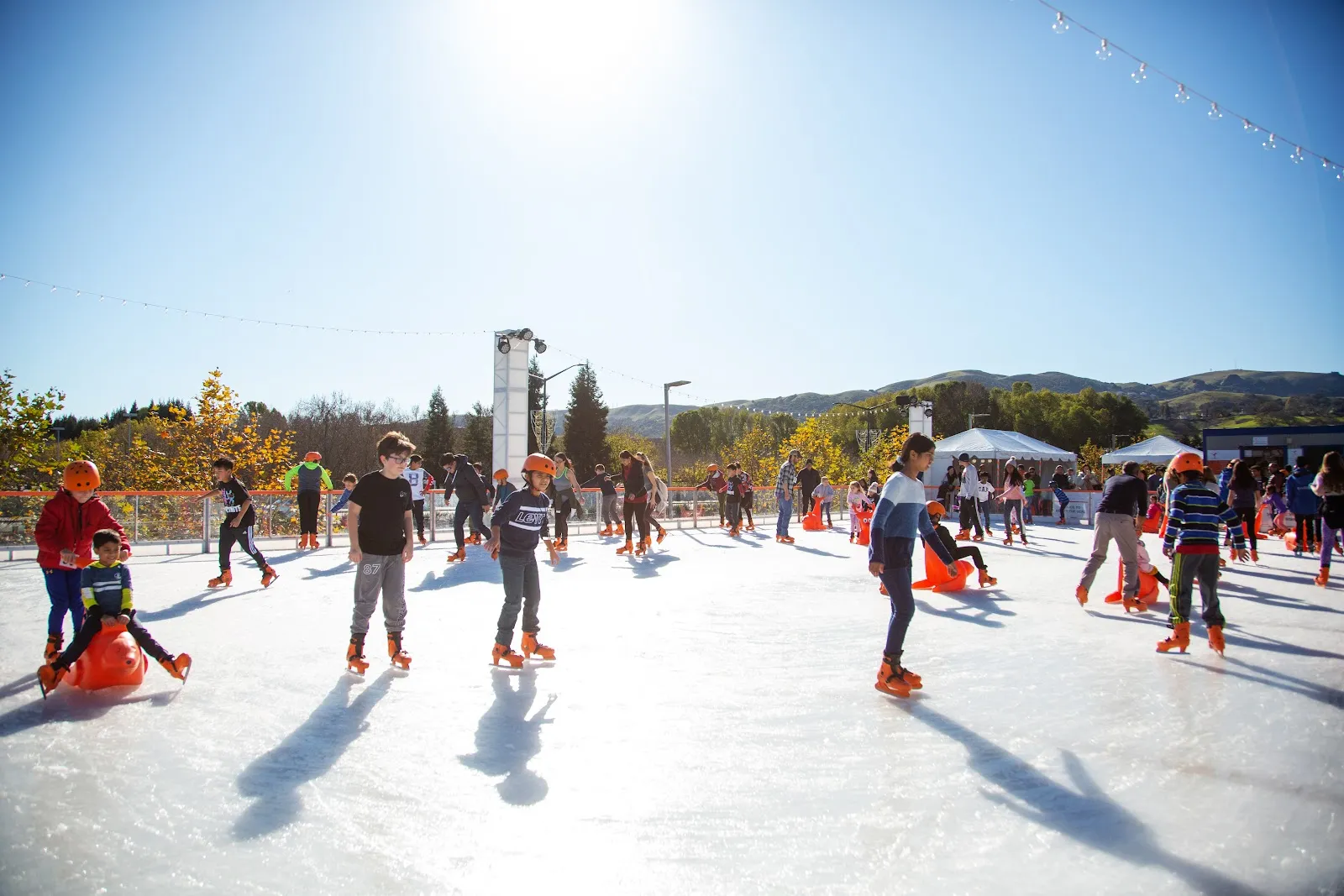 Skaters spread across the sunlit Holiday Ice Rink in San Ramon with long shadows on the ice and hills behind. — Kristi Yamaguchi Holiday Ice Rink
