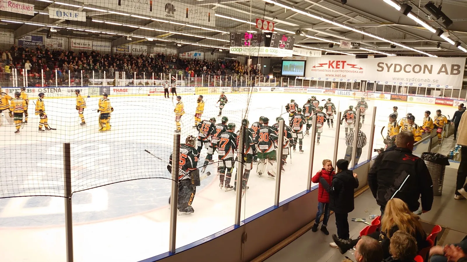 En match på isen i Kristianstad Ishall, med spelare och åskådare längs sargen och sponsorreklam ovanför läktarna. – Ishallen Kristianstad