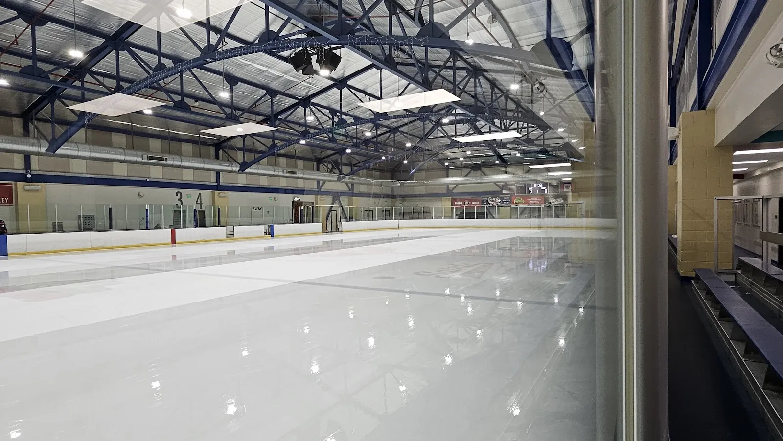 La pista vacía de tamaño NHL vista a través del vidrio junto a las gradas, bajo armaduras de acero azules del techo con tragaluces. — Kroc Center Ice