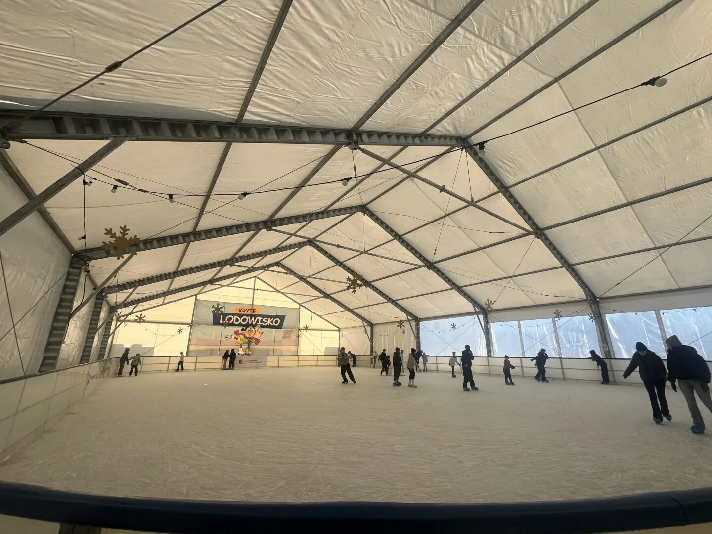 Skaters spread across the boarded ice inside the white steel-framed tent hall of the Wadowice covered rink, with a Kryte Lodowisko banner at the far end and strings of lights under the roof. — Kryte Lodowisko MOSiR Wadowice