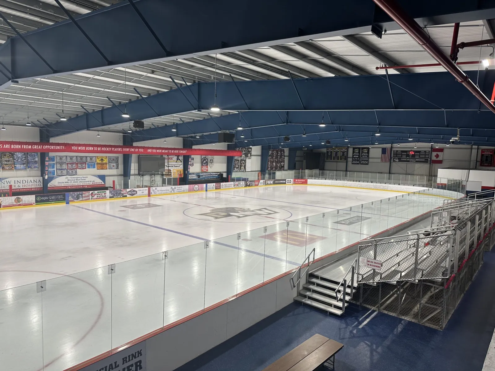 La pista vacía de tamaño NHL vista desde una esquina por encima del vidrio, con gradas de aluminio a la derecha, letreros de patrocinadores y mantas a lo largo de la pared del fondo y armaduras del techo azul oscuro arriba. — KUBE Sports Complex