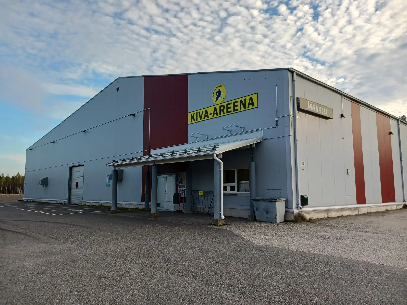 The entrance front of Kuhmo's ice hall, the Kiva-Areena, with its yellow logo sign and the JÄÄHALLI sign on a grey and red steel-clad building under a cloudy sky. – Kuhmon jäähalli