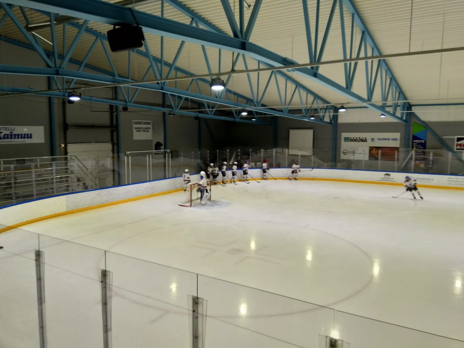 The Kuhmo ice hall with its blue steel arches and a few skaters across the wide ice. – Kuhmon jäähalli