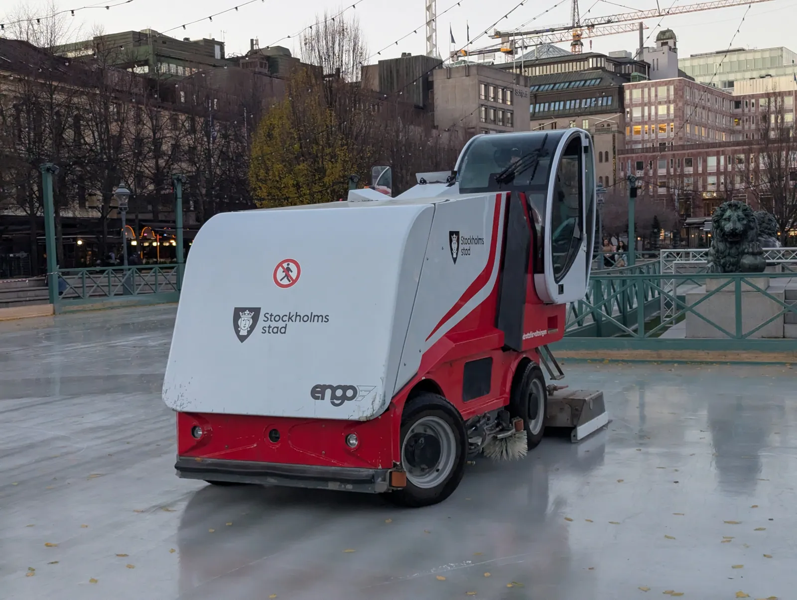 The red and white ice resurfacer on the freshly made ice of Kungsträdgårdens isbana, with the park's green railings, a bronze lion and the Stockholm buildings behind — Kungsträdgårdens isbana