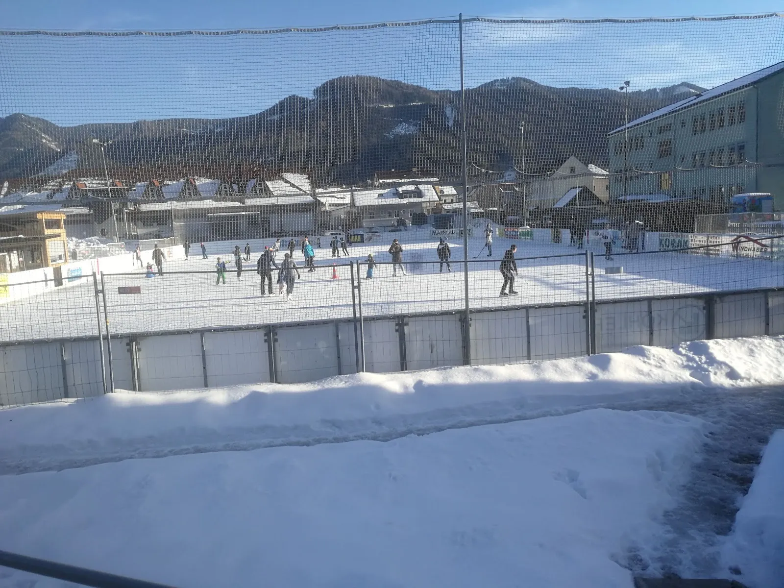 Skaters on the open-air ice of the Kunsteisanlage Langenwang on a sunny winter day, with the mountains and the school behind – Kunsteisanlage Langenwang