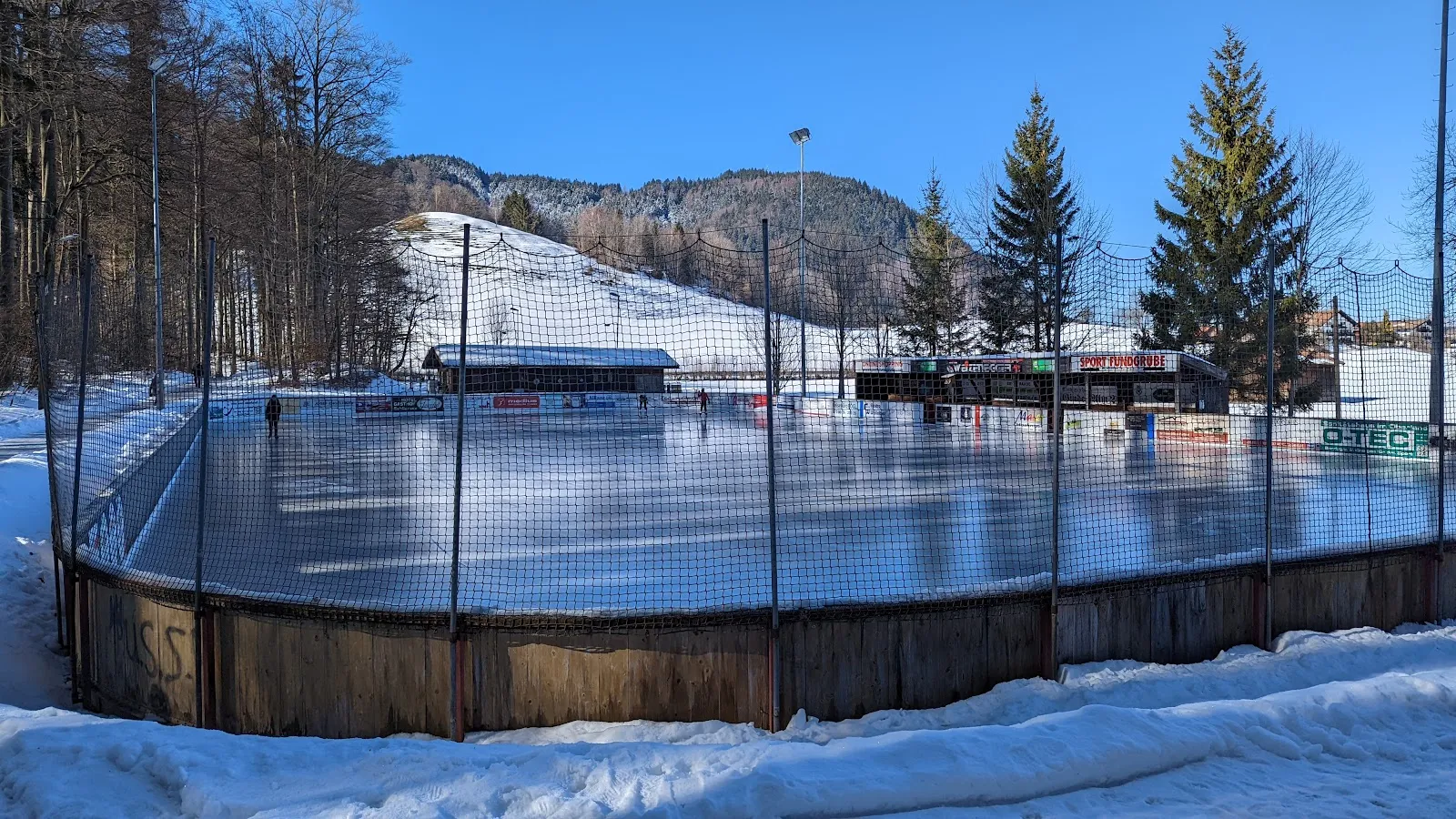 Die Freiluft-Kunsteisarena Tegernsee von außen hinter den Banden mit Holzkante an einem sonnigen Wintertag, mit Schnee am Hang und Tannen dahinter. – KunsteisArena Tegernsee
