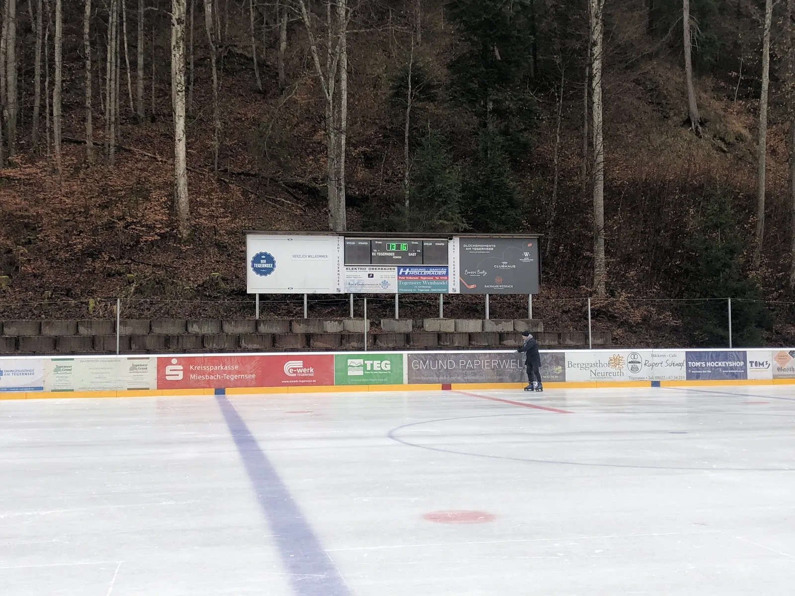 Das leere Freiluft-Eis bei Tageslicht mit einer blauen Mittellinie und einem Anspielpunkt, Werbebanden und einer Anzeigetafel an der gegenüberliegenden Seite vor dem bewaldeten Hang. – KunsteisArena Tegernsee
