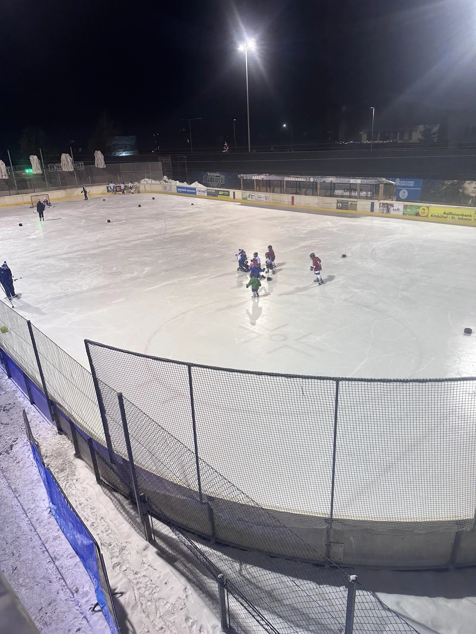 Die beleuchtete Freiluft-Eisfläche der Kunsteisbahn in St. Johann in Tirol bei Nacht, von der Bande aus gesehen, einige kleine Kinder laufen weit hinten auf dem Eis – Kunsteisbahn Panorama Badewelt St. Johann