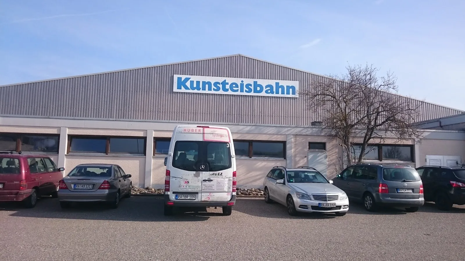 Die lange graue Halle mit dem blauen Schriftzug Kunsteisbahn über dem Eingang und einer Reihe geparkter Autos davor unter blauem Himmel. – Kunsteisbahn Balingen