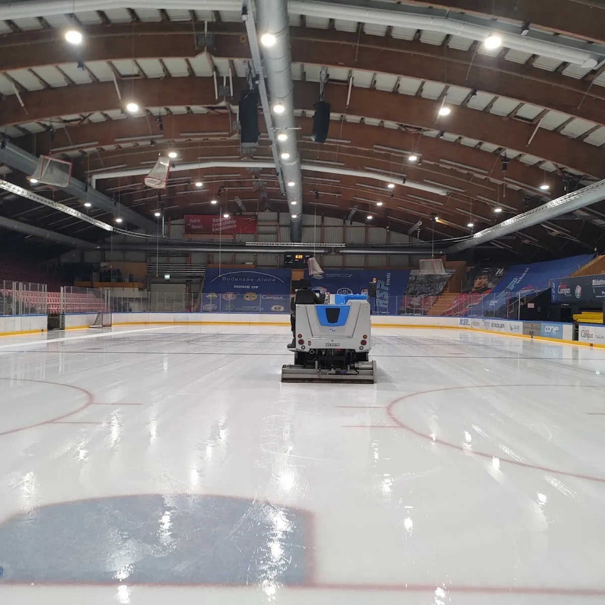 An ice resurfacer crossing the empty ice of the Bodensee Arena under its arched timber roof, with the boards and stands around the rink. — Bodensee Arena
