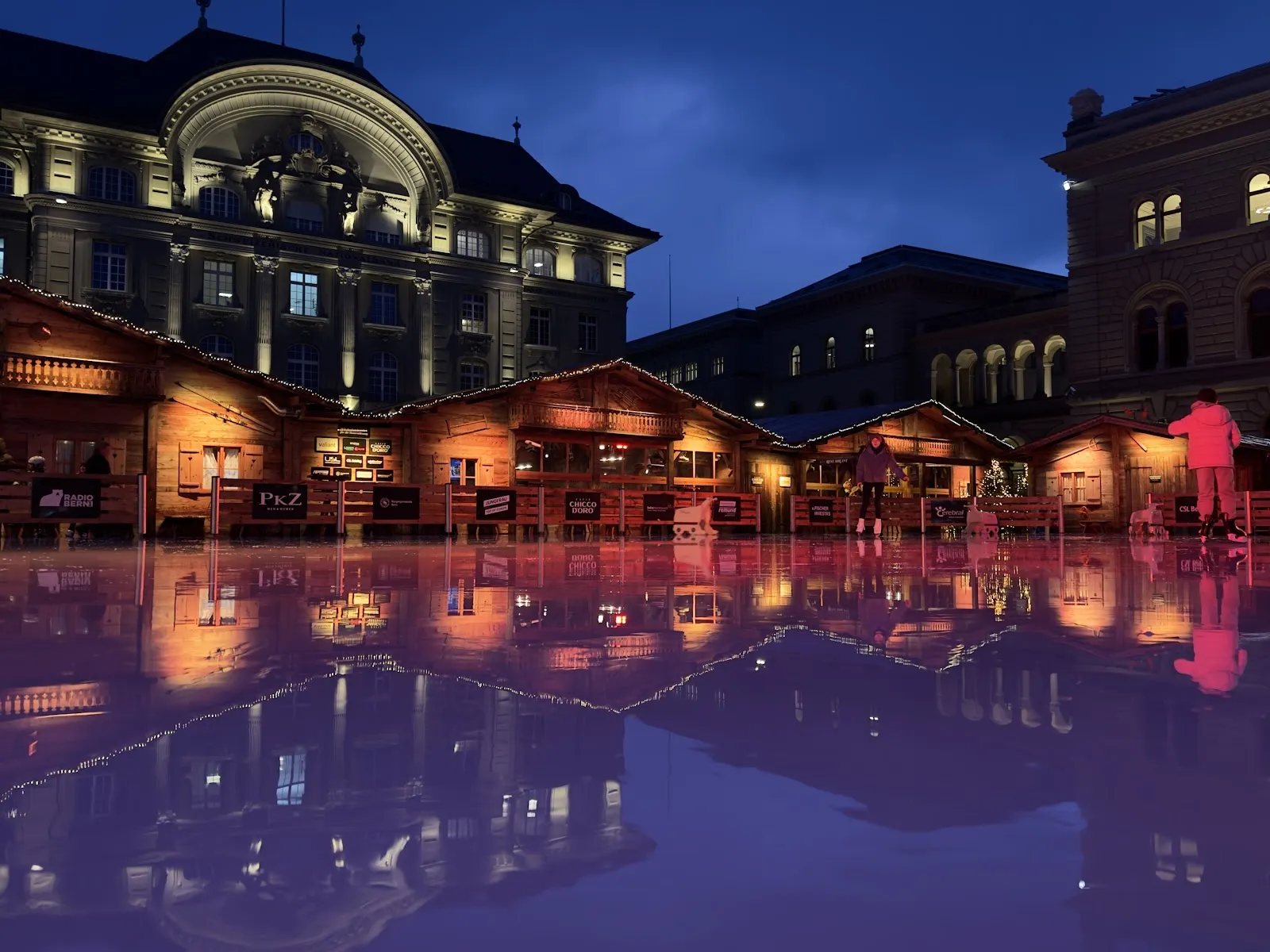 Die Eisbahn auf dem Bundesplatz in Bern in der Abenddämmerung, mit den Holzchalets und den Gebäuden des Bundeshauses, die sich im glatten Eis spiegeln – Kunsteisbahn Bundesplatz