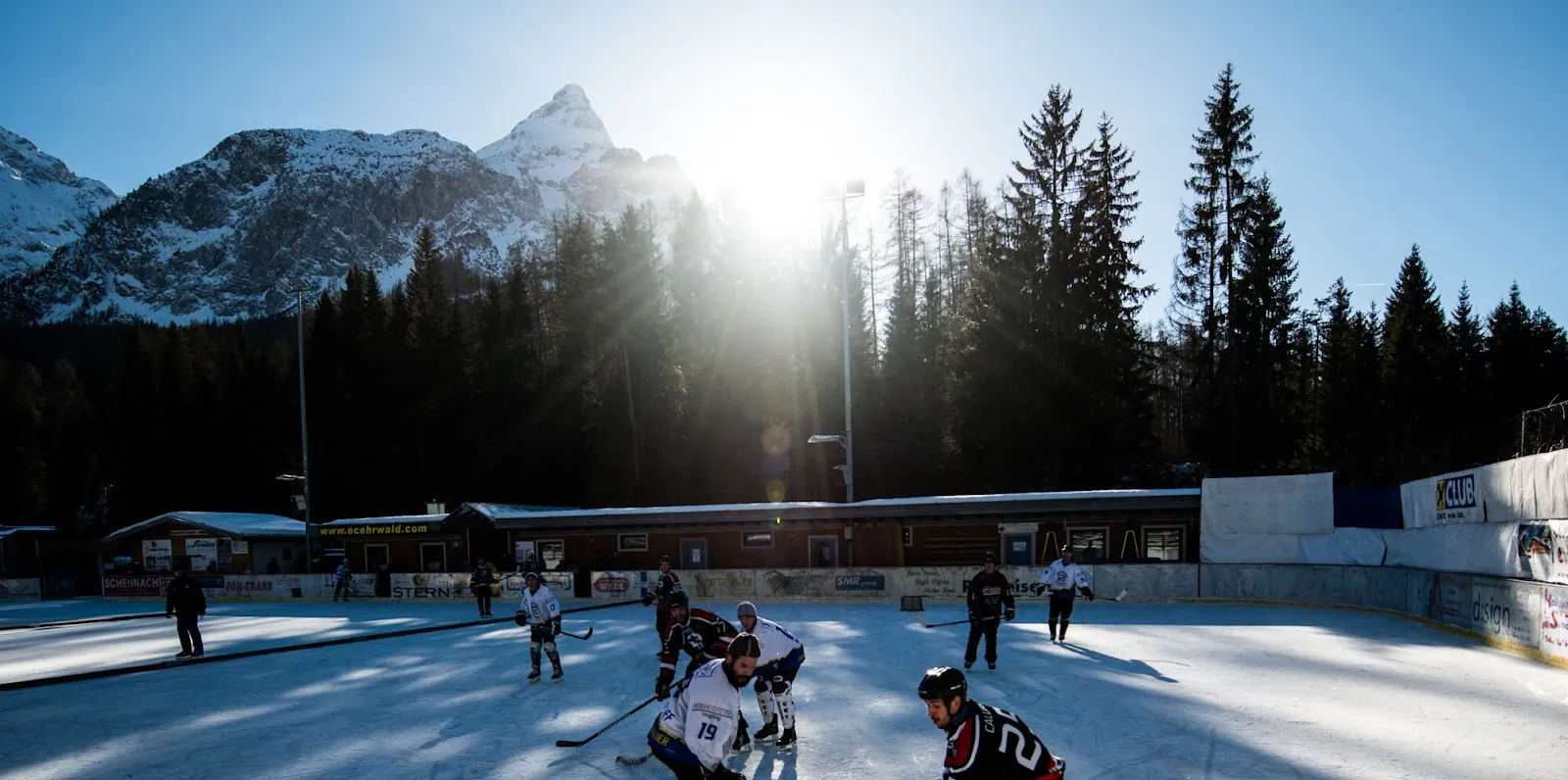 The open-air Kunsteisbahn Ehrwald in winter sun with a hockey game on the ice and the Tyrolean mountains behind. — Kunsteisbahn Ehrwald
