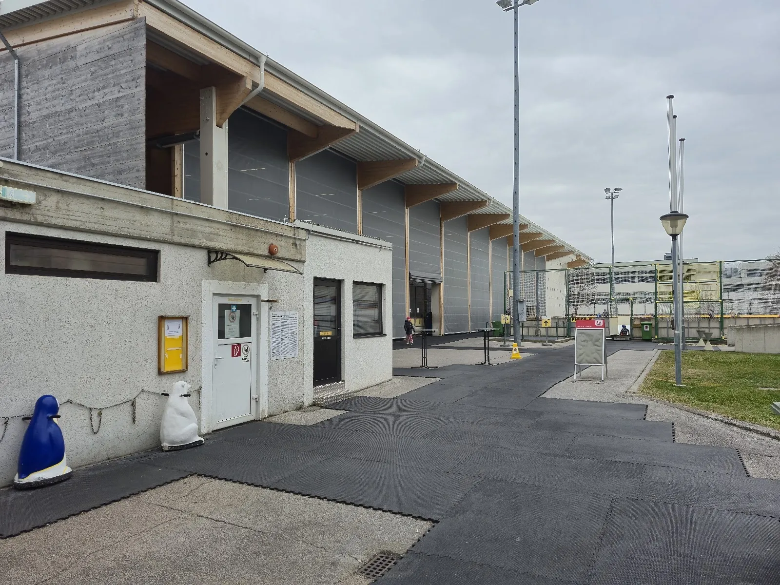 Die Seite der Halle der Kunsteisbahn Eisenstadt vom Zugangsweg aus gesehen, mit der niedrigen hellen Wand und dem Flutlichtmast dahinter. – Kunsteisbahn Eisenstadt