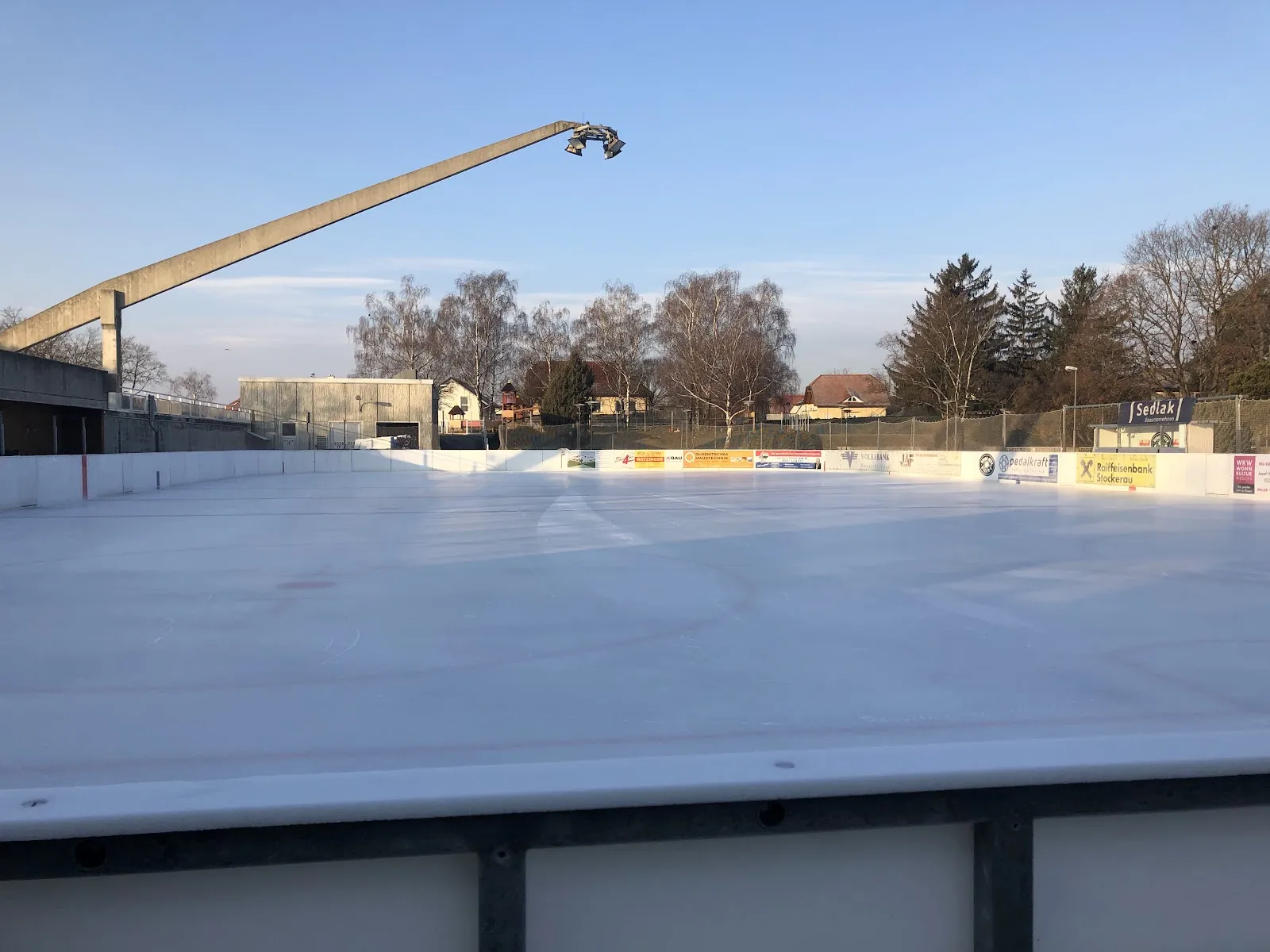 Die leere Freiluft-Eisfläche der Kunsteisbahn Stockerau im Winterlicht, mit den Banden, einem hohen Flutlichtmast und Bäumen dahinter – Kunsteisbahn Erholungszentrum Stockerau