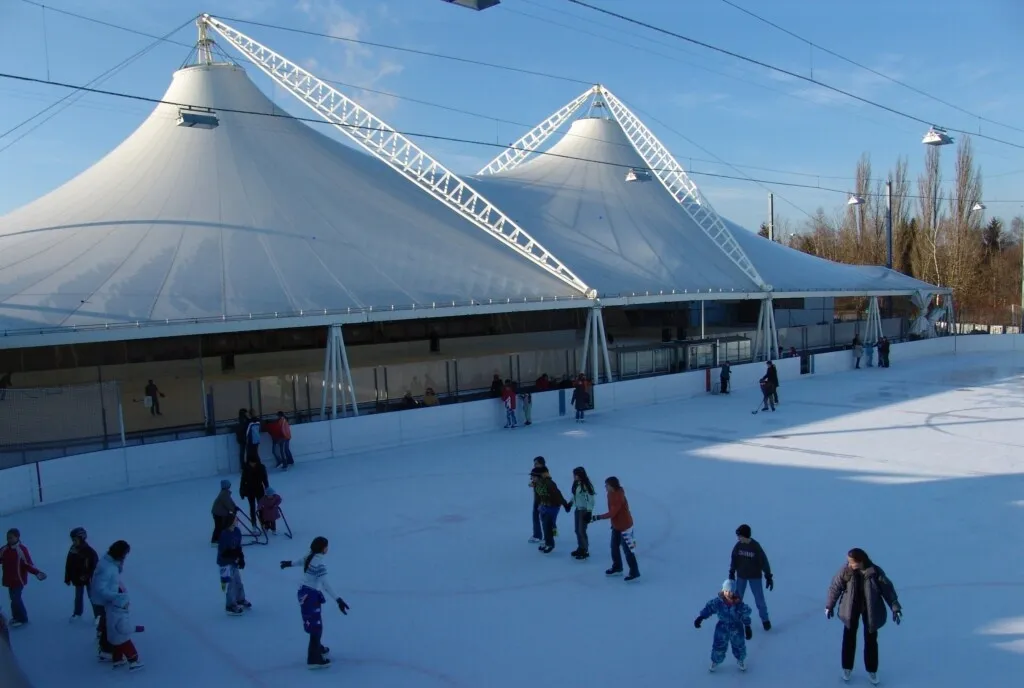 The white tent roofs of the Kunsteisbahn Erlen in Dielsdorf seen from outside on a winter day, with skaters on the ice under the open sides. — Kunsteisbahn Erlen