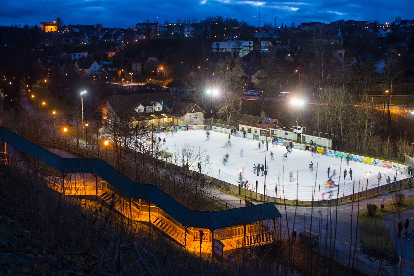 Evening view of the ESV Zwettl artificial ice rink from above, with the footbridge and the floodlit sheet full of skaters — Kunsteisbahn ESV Zwettl