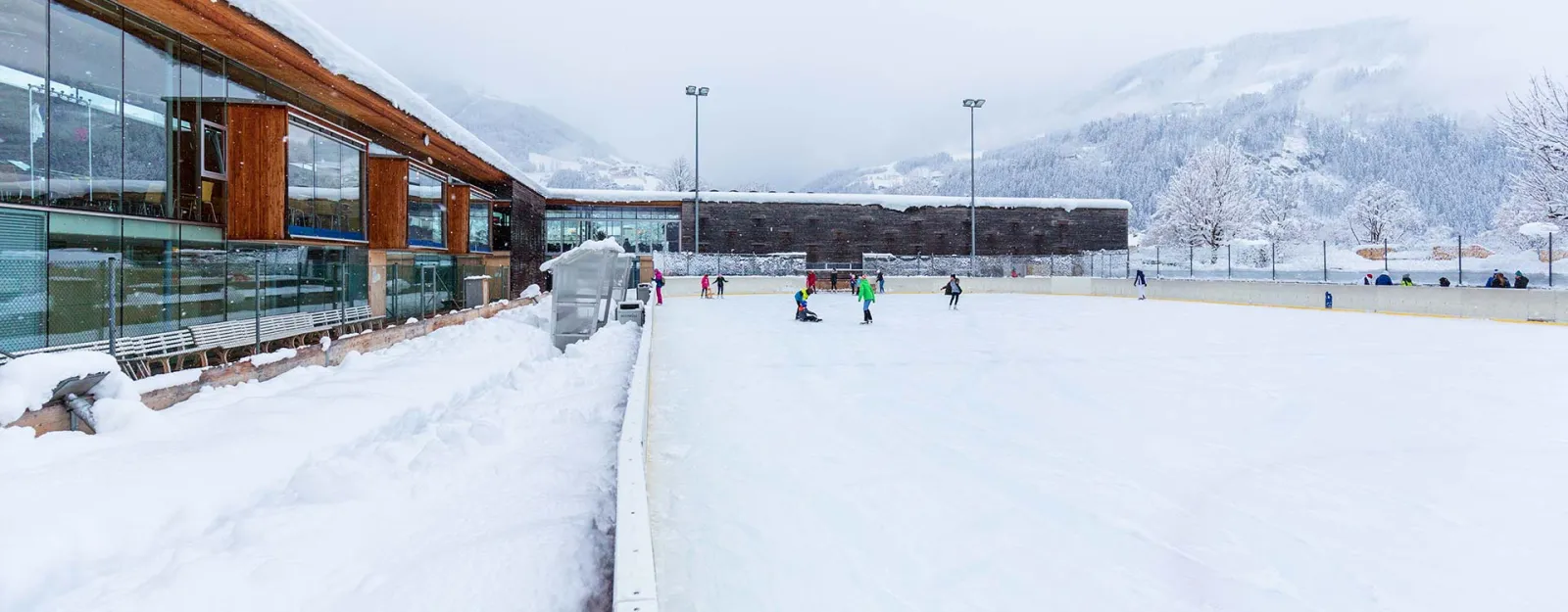 The Freizeitpark Zell artificial ice rink in snow, with the glass-fronted park building on the left, floodlight masts and the Zillertal slopes behind. — Kunsteisbahn Freizeitpark Zell