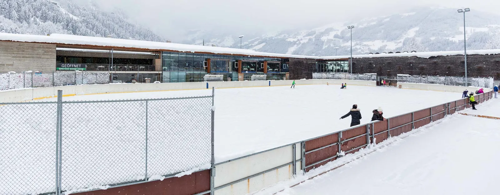 The wide ice surface of the Zell am Ziller rink from the boards, with a few skaters, the park building behind and snowy hills beyond. — Kunsteisbahn Freizeitpark Zell