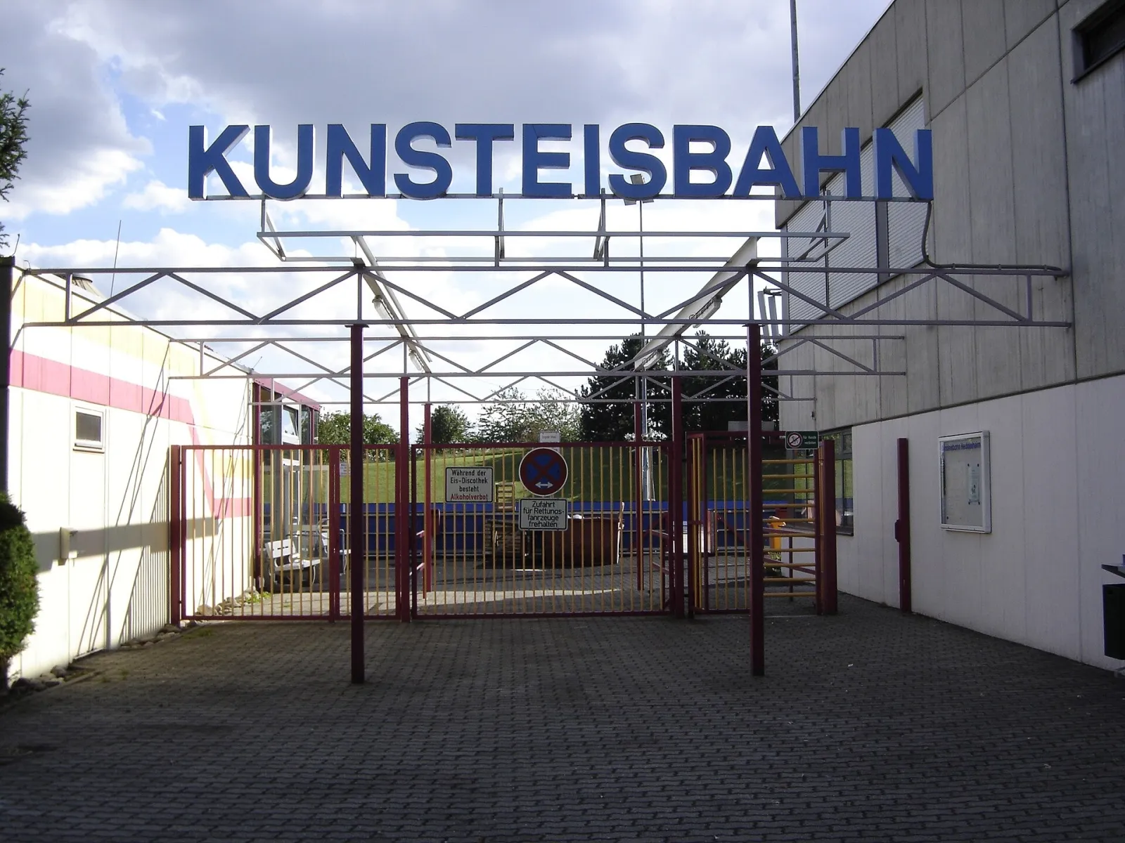 Das Eingangstor der Kunsteisbahn Heddesheim mit dem Schriftzug Kunsteisbahn über dem Stahlrahmen und der Hallenwand daneben. – Kunsteisbahn Heddesheim