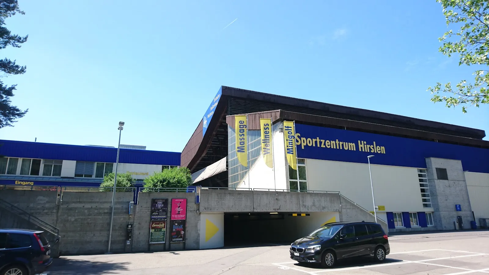 The Sportzentrum Hirslen building in Bülach with its blue name band and sloping timber roof, seen from the car park under a blue sky — Kunsteisbahn Hirslen