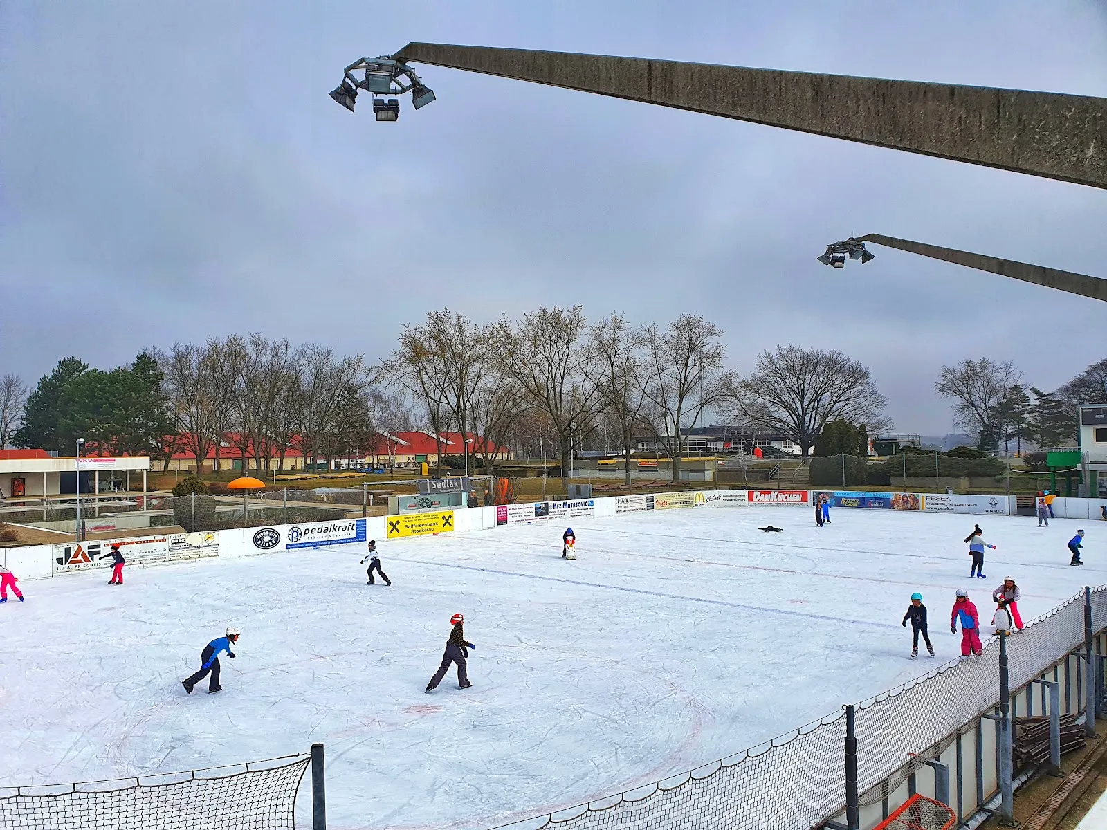 The open-air Kunsteisbahn Hollabrunn seen from the stand, with skaters across the rink and the park trees behind. — Kunsteisbahn Hollabrunn