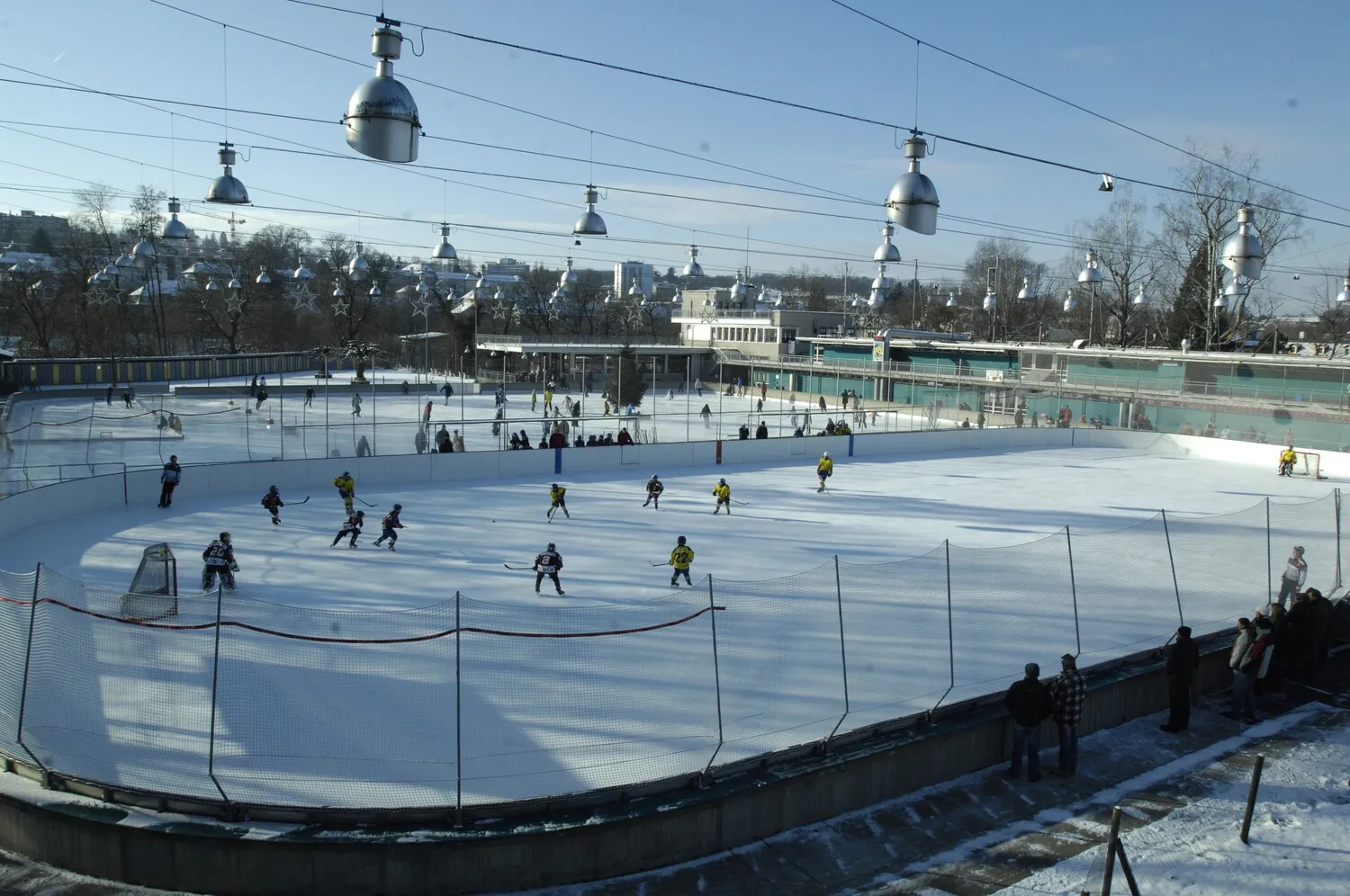 Spieler auf der Freiluft-Kunsteisbahn Ka-We-De in Bern, von der Tribüne neben der Bande aus gesehen, unter den Flutlichtmasten. – Kunsteisbahn Ka-We-De