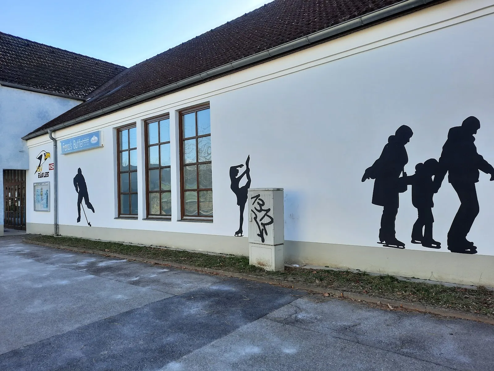 Das weiße Gebäude der Kunsteisbahn Krems mit schwarzen Silhouetten von Eisläuferinnen und Eisläufern und einem Eishockeyspieler, aufgemalt an der Wand neben den hohen Fenstern – Kunsteisbahn Krems