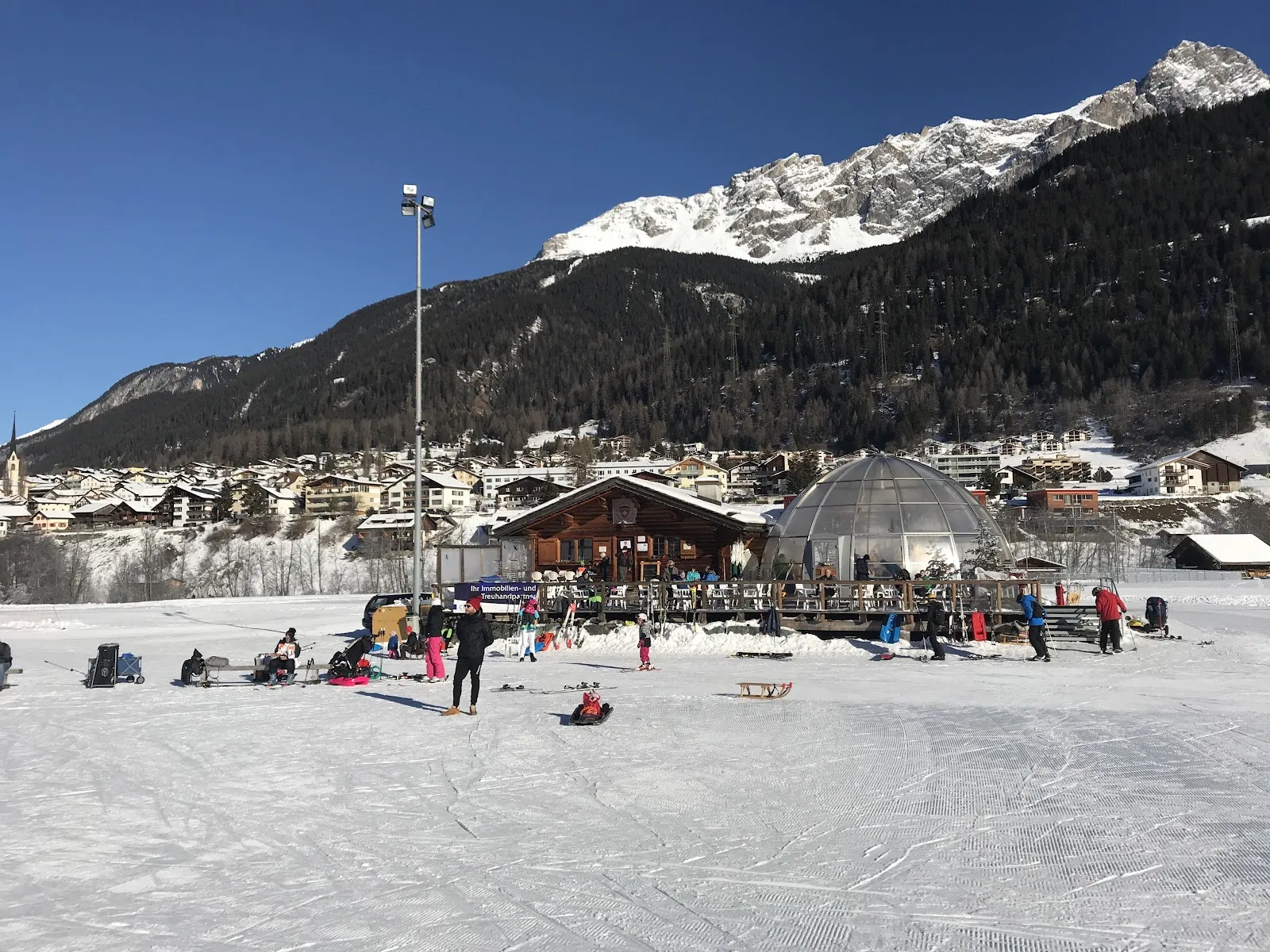 Skaters and families on the ice of the ewz-Arena La Nars in Savognin, with the village beyond — Kunsteisbahn La Nars (ewz-Arena)
